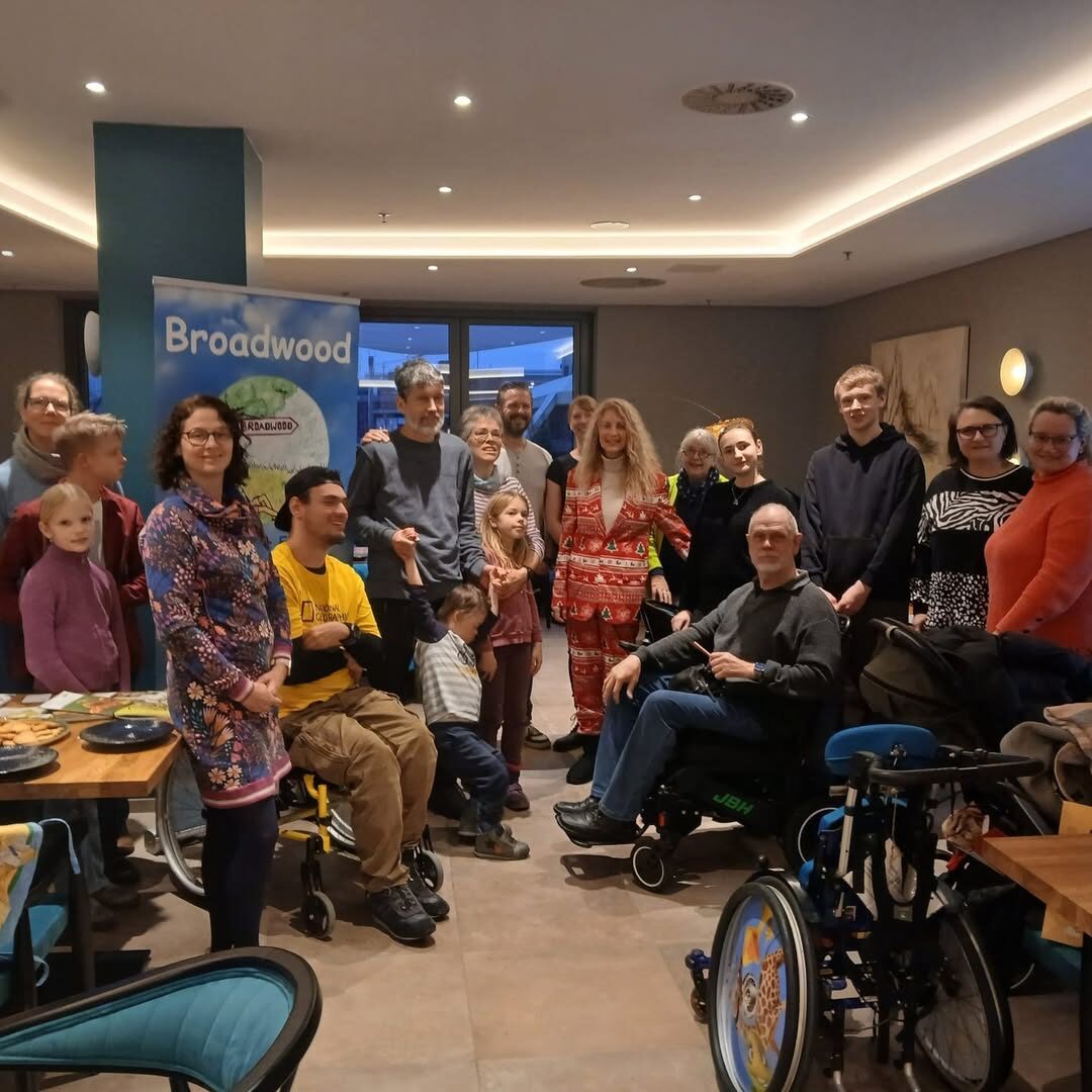 Ein Gruppenfoto. Mehrere Menschen, davon zwei im Rohhlstühl, sitzen und stehen vor einem Roll-Up in einem Restaurant.