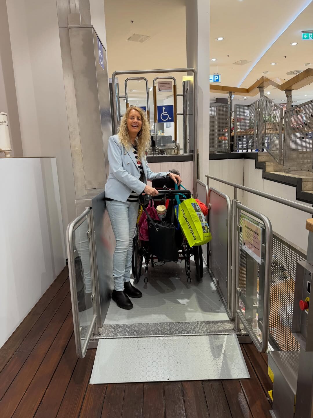 Eine Person steht neben einem Rollstuhl auf einer vertikalen Plattformhebebühne in einem Einkaufszentrum. Die Hebebühne ist mit seitlichen Geländern und einer kleinen Auffahrrampe ausgestattet. Im Hintergrund sind Geschäfte, Geländer und Hinweisschilder für barrierefreie Zugänge zu sehen.