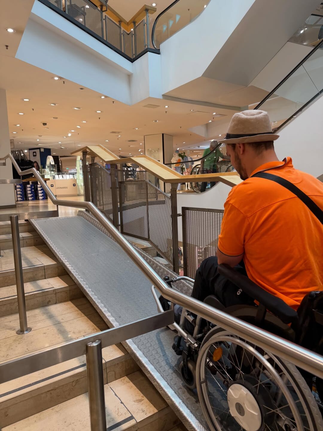 Eine Person im Rollstuhl befindet sich vor einer langen, schrägen Rampe neben einer Treppe in einem mehrstöckigen Einkaufszentrum. Die Rampe führt auf die nächste Ebene. Im Hintergrund sind Verkaufsflächen, Geländer und eine Rolltreppe sichtbar.