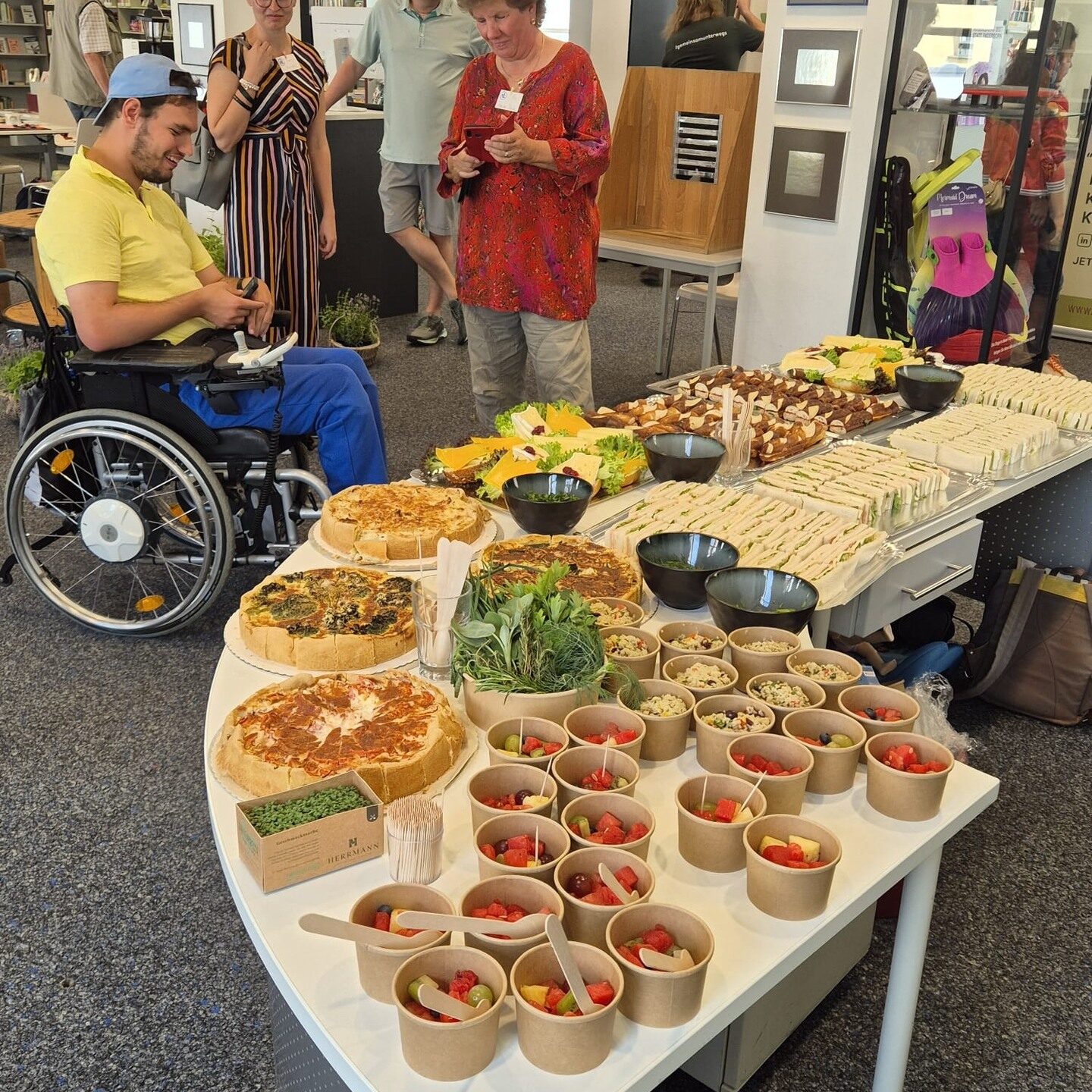 Rechs sitzt Nils, der im Rollstuhl sitzt und das Buffet fotografiert. Daneben stehen weitere Menschen, die sich auf das leckere Essen freuen.