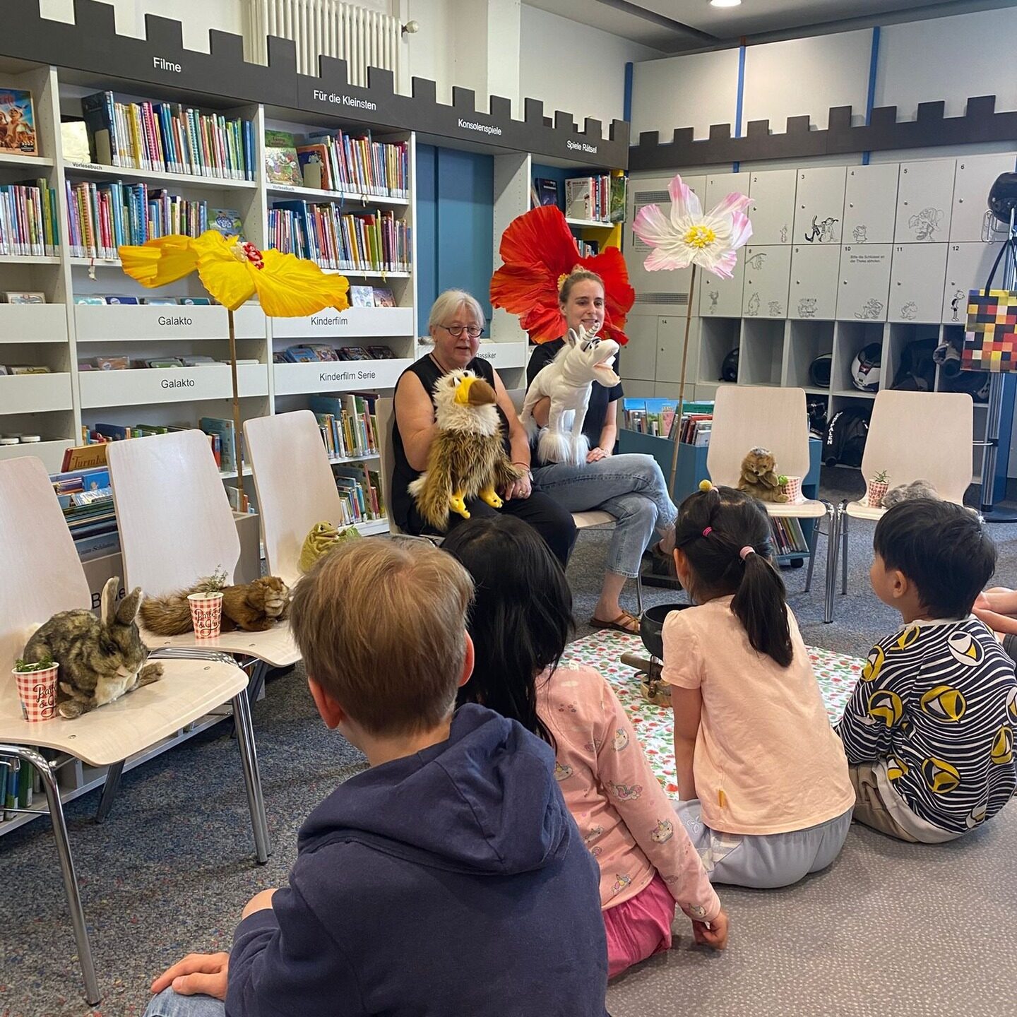 Gisela und Esther stehen hinter Stühlen mit Kuscheltieren. Davor sitzen Kinder und Erwachsene für das Mitmach Theaterstück.