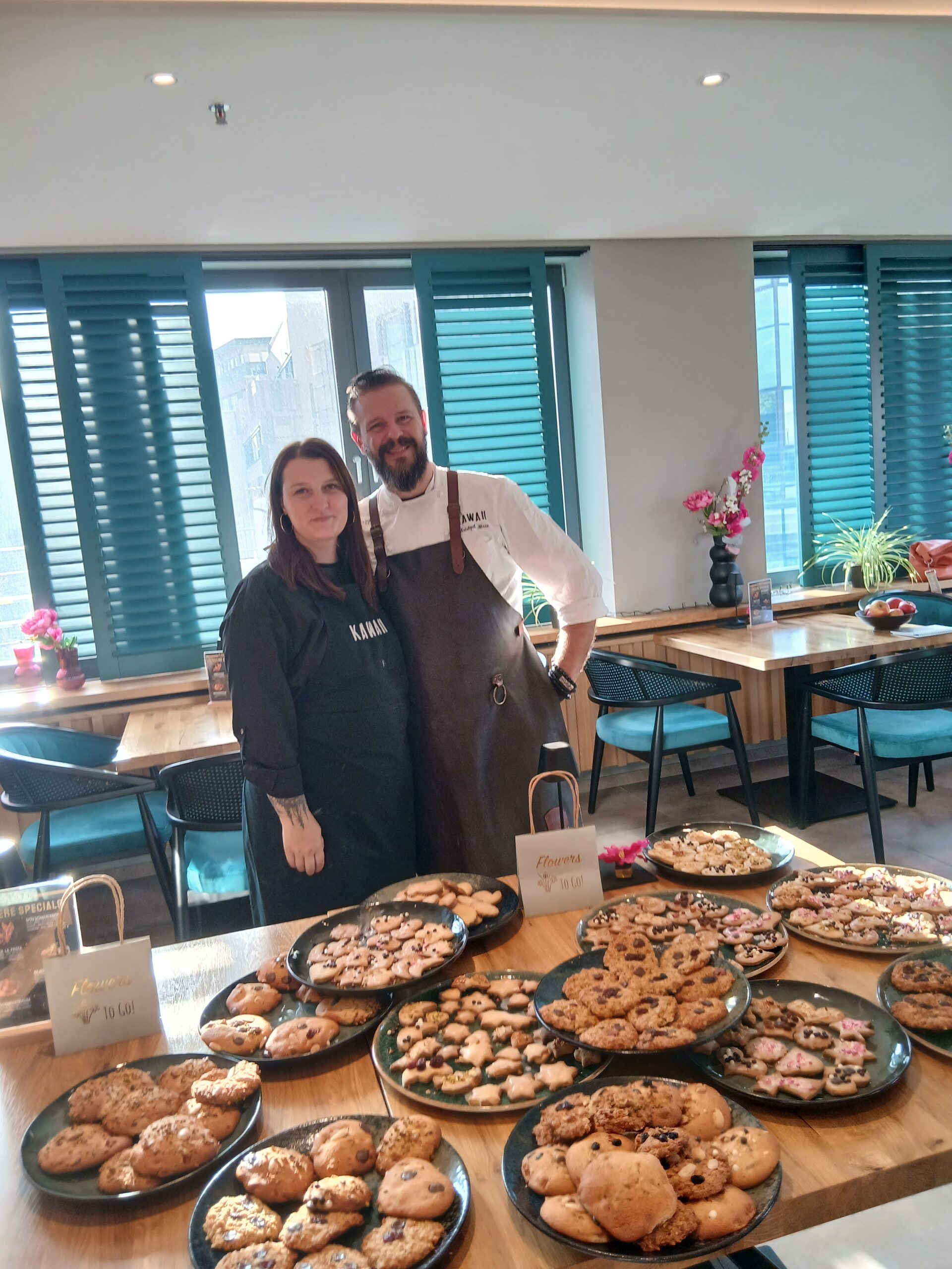 Chefkoch Christoph mit schwarzer Schürze und Assitentin Casandra stehen lächeln vor einem Tisch mit Keksen auf einem Keksbackblech.