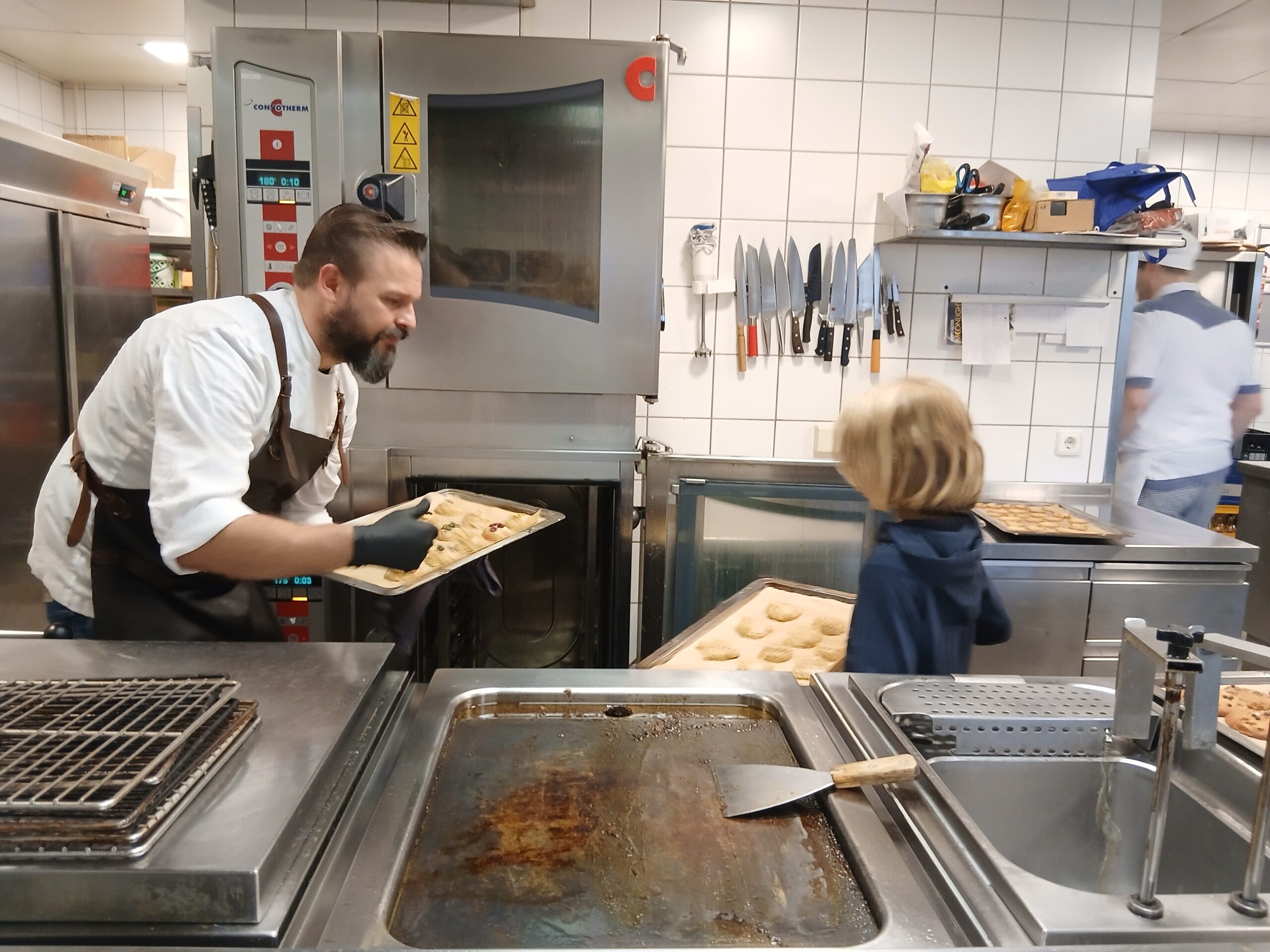 Chefkoch Christoph steht mit einem Jungen in seiner großen Restaurantküche und hält ein Tablet mit Keksen in der Hand. Der Junge hält ebenfalls ein Tablett. Sie schauen sich an.