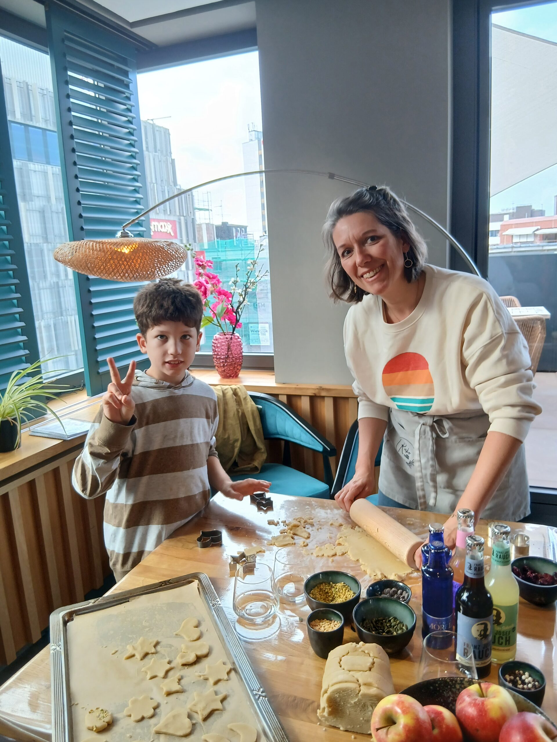 Eine Mutter und ihr Sohn blicken in die Kamera. Der Sohn hat eine Hand zum "Peace"-Zeichen erhoben. Vor ihnen ist ein Tisch mit Backutensilien.