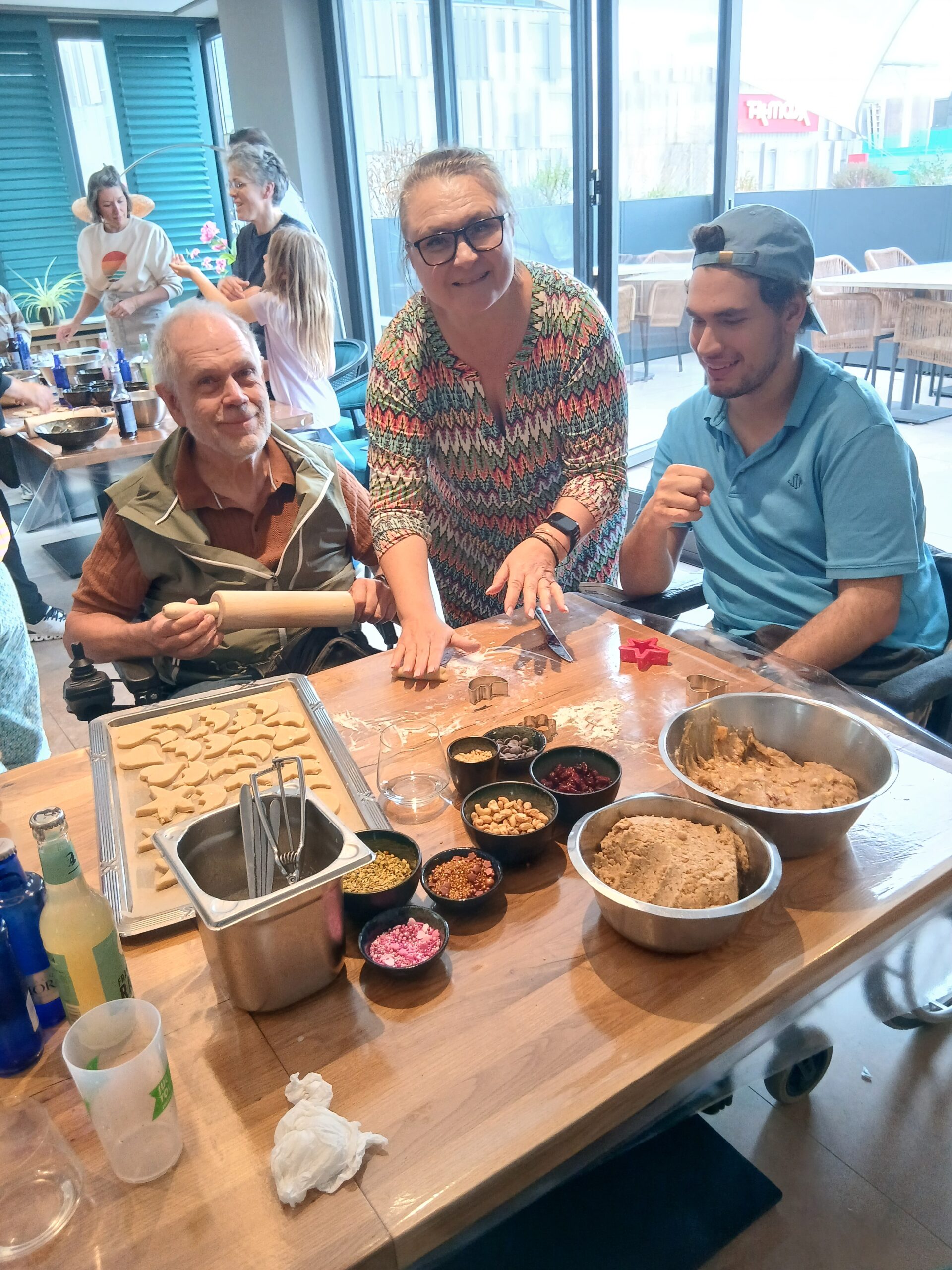 Eine Dreiergruppe von Menschen steht und sitzt um einen Tisch versammelt. Ein Mann im Rollstuhl, eine Frau und ein junge im Rollstühl mit blauer Basecap. Vor ihnen sind Schüsseln mit Teig und Nüssen aufgestellt. Der Tisch ist mit Mehl bedeckt. Links neben ihnen steht ein volles Backblech mit herzförmigen Keksen. Die Frau verteilt bunte Keksförmchen in verschiedenen Formen.