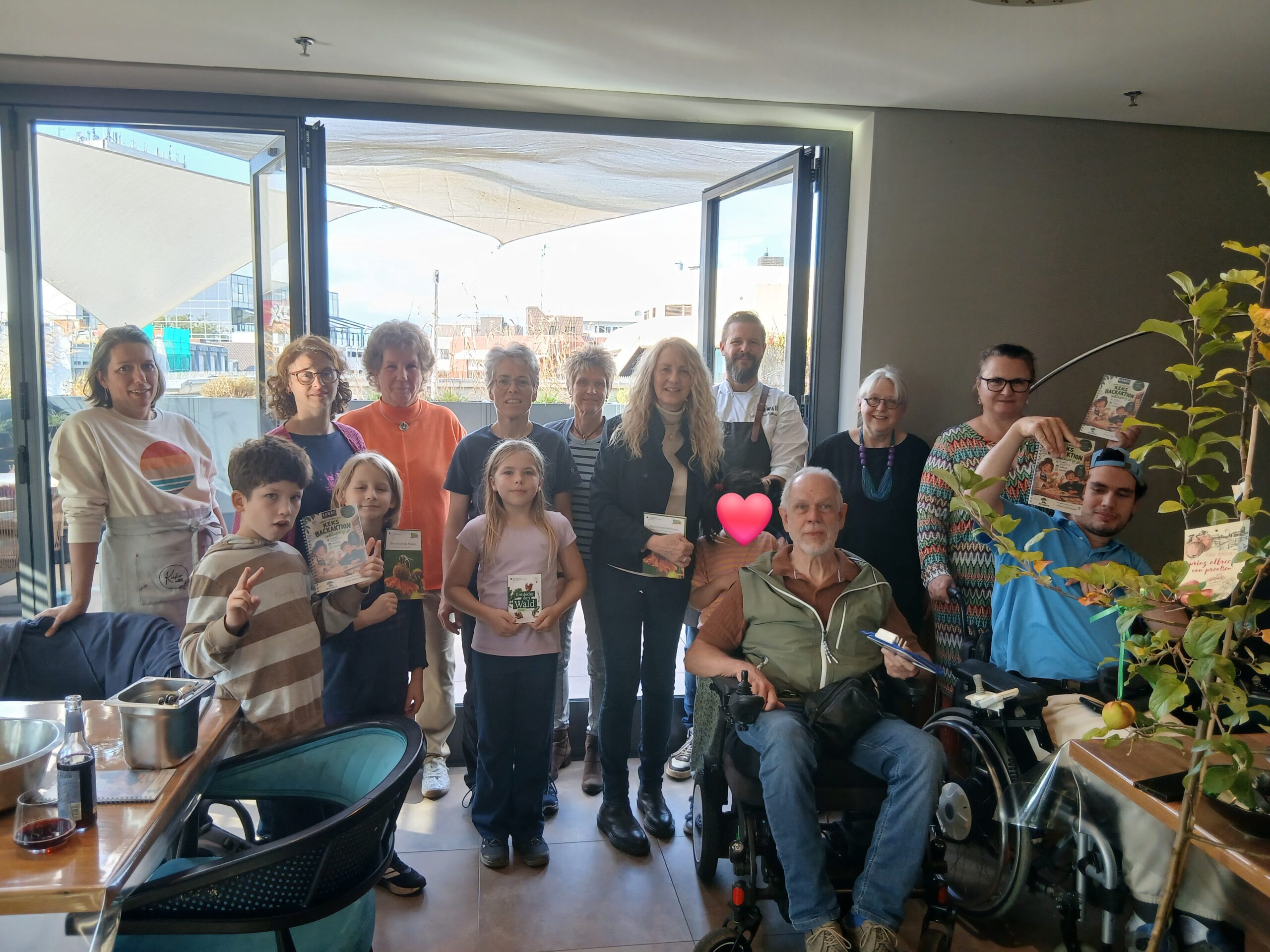Eine inklusive Gruppe mit Kindern und Erwachsenen und Geschäftsführer und Koch Christoph Hesse steht vor der Terrassentür im Kawaii Restaurant, Re. daneben steht ein Apfelbäumchen