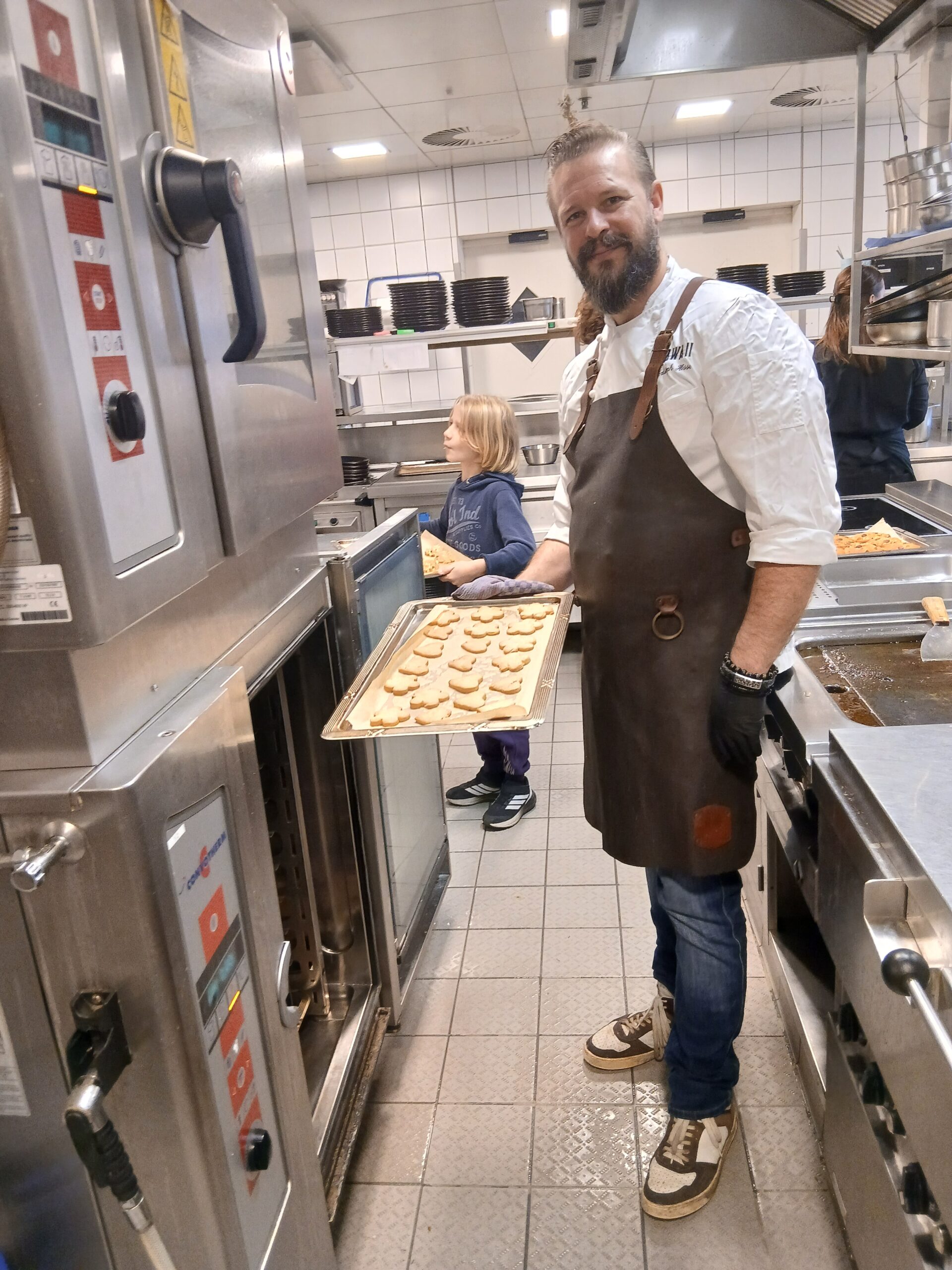 Chefkoch Christoph mit schwarzer Schürze steht vor einem großen Ofen mit einem Backblech voller herzförmiger Kekse in der Hand. Neben ihm steht ein Junge und blickt neugierig in einen zweiten Ofen.
